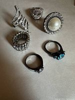 Photo shows six assorted rings on a neutral surface, featuring various stones and metal finishes.