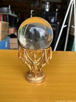 Crystal ball on gold statuette holder on a wooden surface with background showing partially a room with various items.
