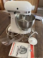 White KitchenAid Classic stand mixer with stainless steel bowl, wire whisk, flat beater, dough hook, pouring shield and manual laid out on wooden table