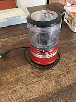 Side view of the small red KitchenAid food processor/blender plugged in, showing the red base and clear blending container on a wooden table.