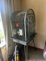 Full view of large metal arched bird cage with wheel base placed in corner near window. Shows perches, food/water containers, and cage door.