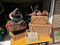 Photo shows eight assorted wicker baskets placed on and around a yellow table outdoors. Includes picnic basket with leather straps, small handled basket, decorative fabric lined basket, two conical hanging baskets, and other woven baskets.