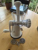 Front angled view of manual meat grinder showing white body, clear plastic hopper, white handle, suction base, and two metal grinding plates with sharpening blades placed in front.