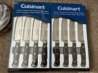 Two unopened boxed sets of 4 Cuisinart forged steak knives each, showing front of packaging with knives visible through plastic window