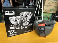 Photo shows boxed set of 12-piece metal mixing and measuring bowls and a stoneware mortar & pestle with label on wooden table.