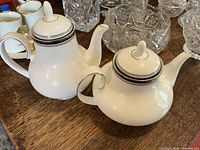 Two Royal Doulton Sarabande pattern white bone china tea pots with black and silver banding on wooden table.