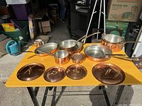 Overhead view of all copper cookware on table, showing five pots/pans, four lids, and mesh strainer
