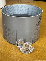 Clear crystal bird figurine with faceted body and black eyes in front of the original silver Swarovski box.