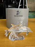 Photo shows clear Swarovski crystal dolphin figurine on a wooden surface with the original silver-grey Swarovski box behind it. The box features the black swan logo and 'Swarovski Silver Crystal' text in white.