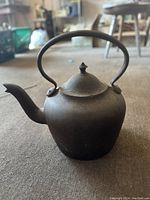 Rounded dark cast iron kettle with domed lid and curved handle taken on floor surface.