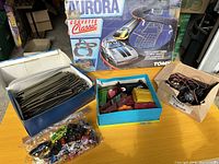 Main photo showing boxed Aurora Corvette Classic set at rear, blue and white box of model train tracks at left, blue accessory box and clear bag of diecast cars center, and cardboard box of wiring at right