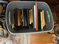 Top-down view inside large grey plastic bin showing multiple picture frames stacked upright, some with wood and metal detailing.