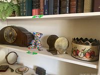 Shelf showing wooden mantle clock, metal cat-shaped basket, brass shell bookends, and a decorative tin basket with apple design.