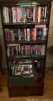 Black bookcase filled with assorted DVD cases of TV shows and movies, with a green woven basket on top holding additional DVDs.