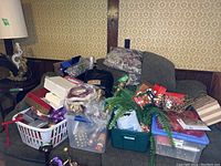 Photo showing several plastic bins filled with Christmas ornaments, assorted boxes, and wrapping bags stacked on a green armchair and sofa around it.