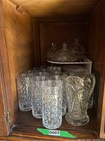 Wide view of cabinet containing multiple crystal highball glasses and crystal butter dishes with lids, plus a large crystal pitcher or vase, showing lot contents and arrangement.
