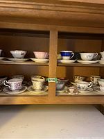 Display of multiple miniature tea cups and saucers arranged on wooden cabinet shelf, showcasing variety of floral and decorative designs, colors include blue, pink, gold, and white patterns.
