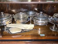 Display of several silver casserole dishes with lids and a silver brush on a wood shelf