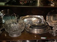 Photo showing various silver serving trays and a glass bowl with etched floral designs and gold trim arranged on a shelf.