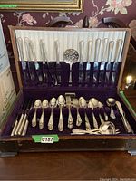 Full view of the antique tarnish proof flatware set inside a wooden case with purple velvet lining showing knives, forks, spoons and serving utensils.