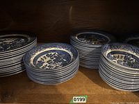 Four stacks of blue and white Blue Willow dinner plates displayed in a wooden cabinet shelf.