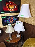 Photo showing the three lamps: a tall metal floor lamp with white shade, two table lamps on a wooden side table; one porcelain Victorian style with painted details and a white shade, the other wood and glass with beige shade.