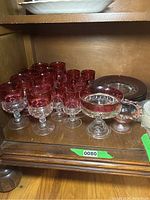 Multiple cranberry rimmed stemware glasses, compote dish and plates on wooden shelf