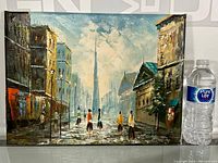 Full front view of signed impressionist oil on canvas painting showing urban street scene, city buildings, figures, and distant spire. Dimensions roughly 16x12 inches as indicated by comparison with water bottle.