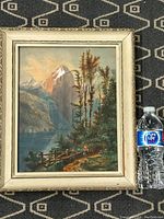 Framed watercolour of mountain landscape with trees, woman, and lake, shown with water bottle for scale