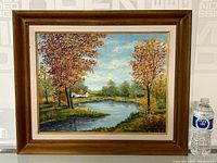 Framed oil painting showing a pond surrounded by autumn trees and two white cottages in the background.