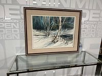 Framed watercolour painting of birch trees against a winter snowy landscape seen from a distance on a glass table.