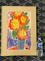 Full view of framed watercolour painting featuring orange flowers with green leaves in a blue vase, positioned on a patterned carpet with a water bottle for size reference.