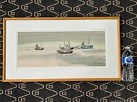 Full view of framed watercolour painting showing multiple fishing boats on calm water with a light wooden frame and white mat.
