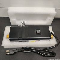 Photo of the compact black water heater unit with power cord and brass connectors, placed on a foam block.