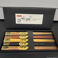 Top view of 5 pairs of Japanese reusable chopsticks arranged in a black presentation box. Each pair is labeled with Japanese text and differs in wood tone and finish.