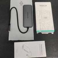 Photo showing the MOKiN 7-in-1 hub with attached USB Type-C cable, product manual, and packaging box, all brand new and unused.
