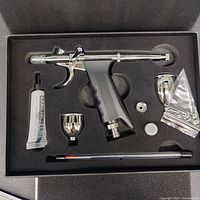 Top down view of the airbrush tool set featuring the airbrush gun, two fluid cups, lubricating oil tube, and small metallic parts neatly fitted into foam inside a black case.