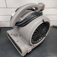 Side angle showing gray plastic body, carrying handle with wrapped power cord, and black front air vent for air output.