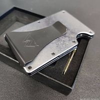 Oblique front view of the slim minimalist metal wallet with black steel money clip, screws visible, inside a black foam-lined box with included screwdriver.