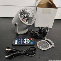 Photo showing the LED projector light assembly, remote control, USB cable, suction cup mount, and additional diffuser cover, all laid out in front of a white box.