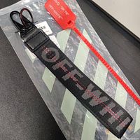 Black Off-White branded braided nylon keychain with a black metal clip and attached signature red plastic zip tie, placed on Off-White clear plastic packaging with diagonal stripes.