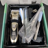 Photo showing glossy black tray inside box with hair clipper, metal file, blue and white handled scissors in plastic sheath, comb, and charging adapter
