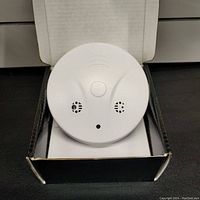 Front view of white round smoke detector device in box showing vents and camera lens.