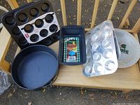 Photo showing all seven baking pans including two aluminum mini muffin pans, two Baker's Secret mini muffin pans, an unused non-stick loaf pan, a used Wolfgang Puck springform pan, and a pie plate arranged on a wooden surface.