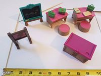 Photo showing pink teacher and student desks with green lunch pails, books, stools, a small and large chalkboard with printed drawings