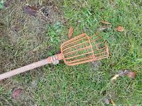 Close-up image of orange wire basket end of fruit picker and wooden pole handle lying on grass.