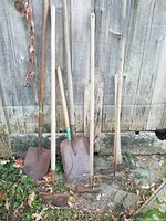 Photo showing seven worn garden tools including shovels, rakes, and hoes leaning against a wooden fence with rusted blades and metal heads.