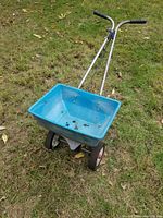 Full view of blue plastic lawn spreader with metal frame, two wheels, and handlebar on grass.