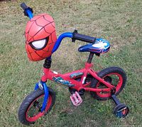 Full view of 12-inch Huffy Spider-Man bike on grass showing red and blue frame, training wheels, Spider-Man mask storage on handlebars, and Spider-Man themed colors and designs.