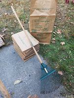 Photo showing a stack of 30 brown paper leaf bags alongside a garden rake with a green metal head and a long ash wood handle labeled 'Arcadia Quality Ash Handle'. The bags are labeled for yard waste like grass, leaves, and garden trimmings.